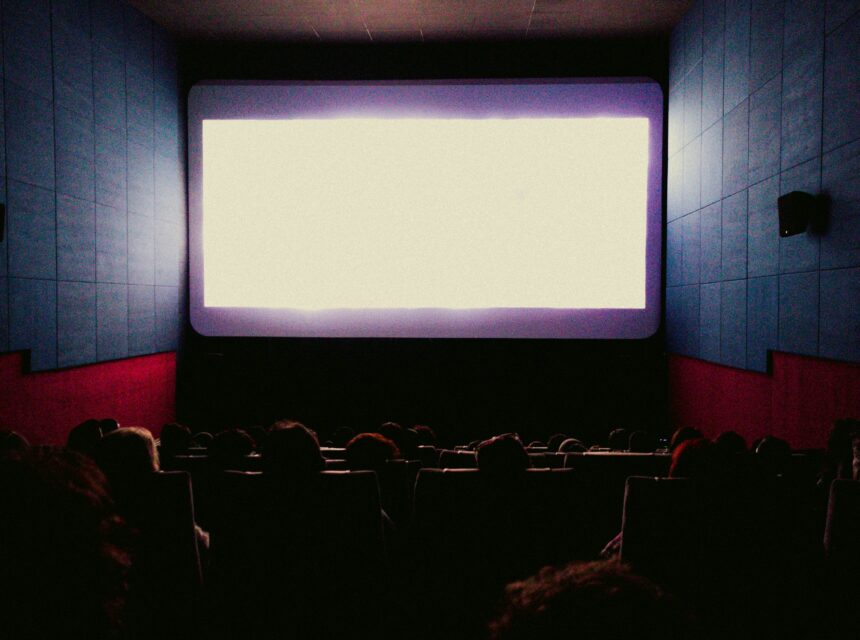 A dimly lit cinema filled with people seated and facing a large, bright white screen, waiting for the film to start.