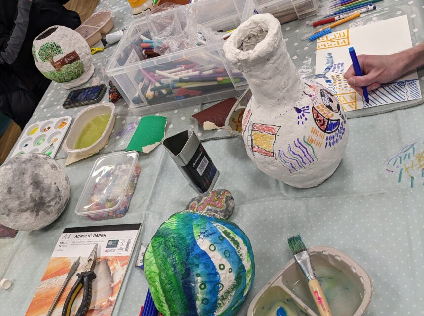 A group of people decorate papier mâché globes and vases at a craft table covered with art supplies, paint, and brushes. Some pieces feature colourful designs, landscapes, and faces.