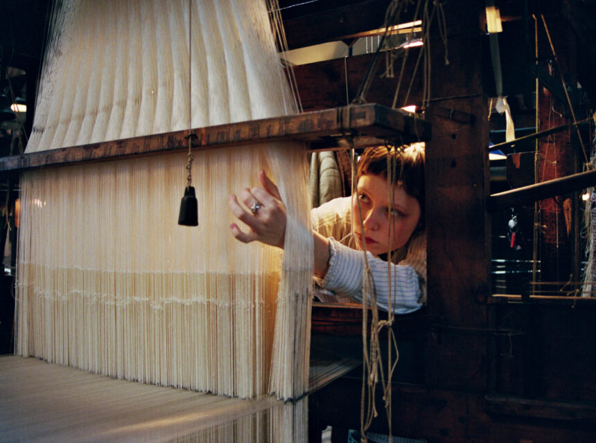 A person carefully adjusts threads on a loom, surrounded by wooden machinery, focused intently on weaving white fibres.