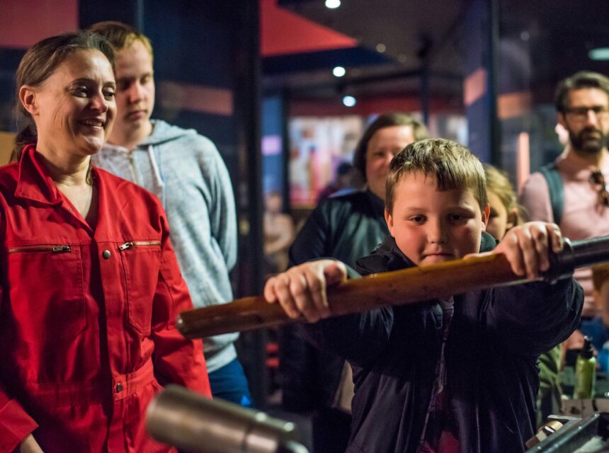 A young boy turns a wooden handle whilst several adults watch and smile in a museum or interactive exhibition setting.