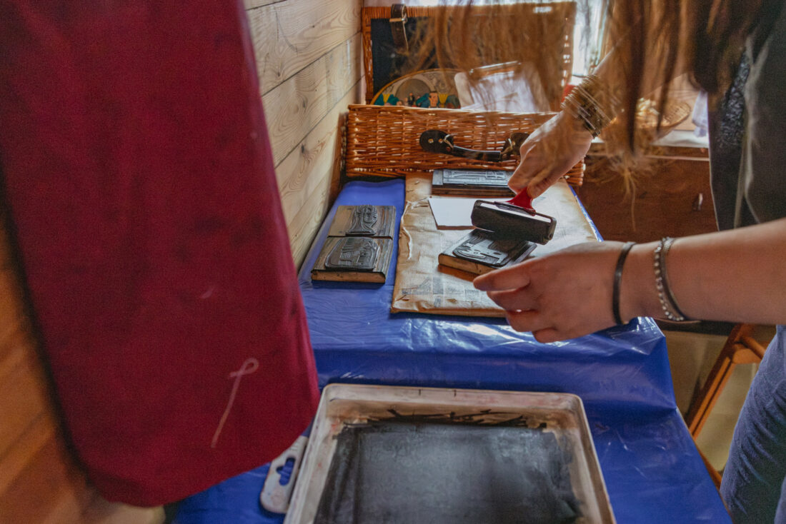 A person rolls ink onto a block for printing, surrounded by tools on a blue-covered table in a cosy, wood-panelled room.