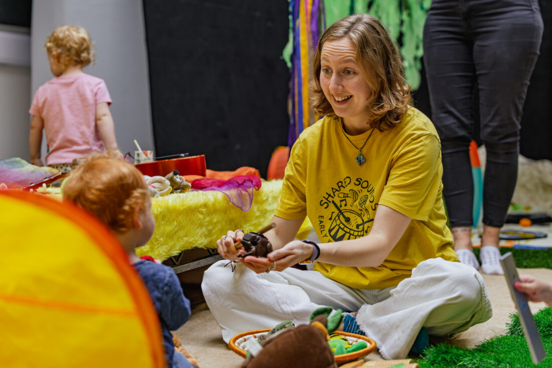 A woman in a yellow shirt sits cross-legged, smiling and holding a small animal, engaging with a young child in a colourful play area. Other adults and children are in the background.