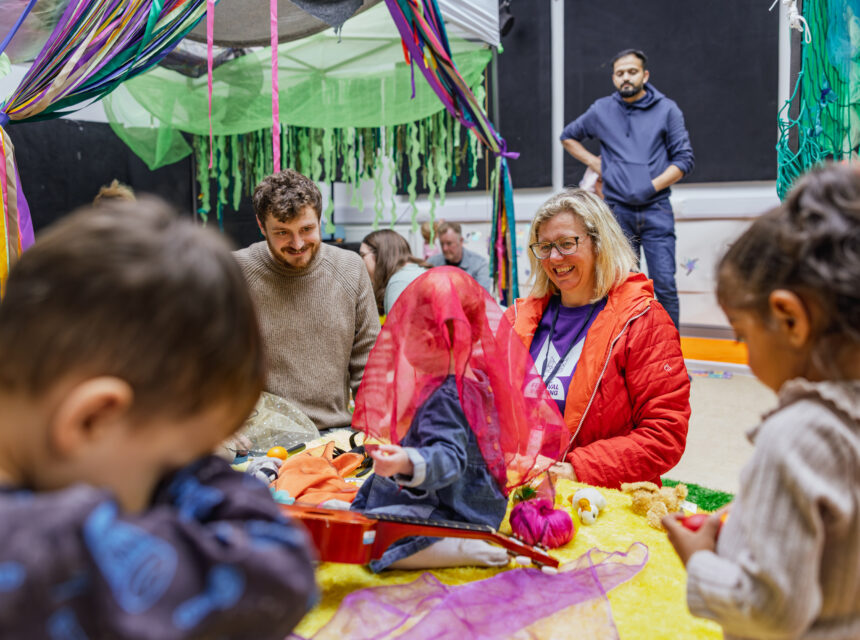 Several adults and young children sit and play together with colourful fabric and toys in a decorated indoor space. One adult smiles while children explore objects. Bright streamers and netting hang around them.