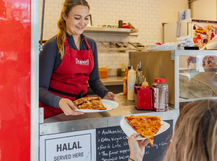 A woman in a red apron serves a slice of pizza on a plate to a customer at a food stall with a Halal served here sign. The scene is bright and welcoming.