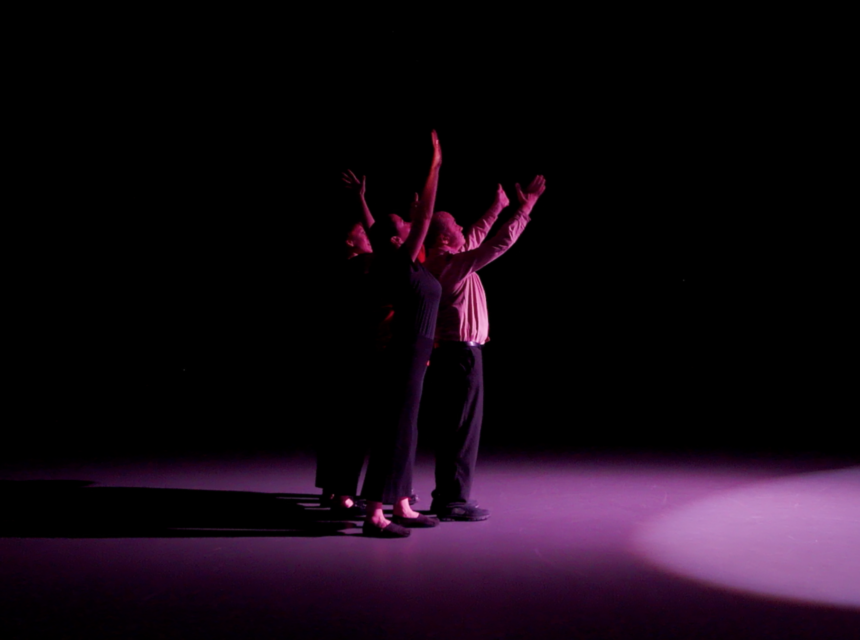 Two people stand close together under a spotlight on a dark stage, reaching upwards with arms outstretched. Their shadows and the edge of the spotlight are visible on the floor.