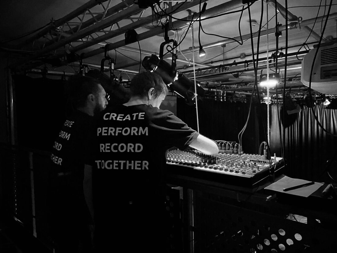 Two people in dark shirts reading CREATE PERFORM RECORD TOGETHER operate an audio mixing desk in a dimly lit backstage area filled with cables and lights.