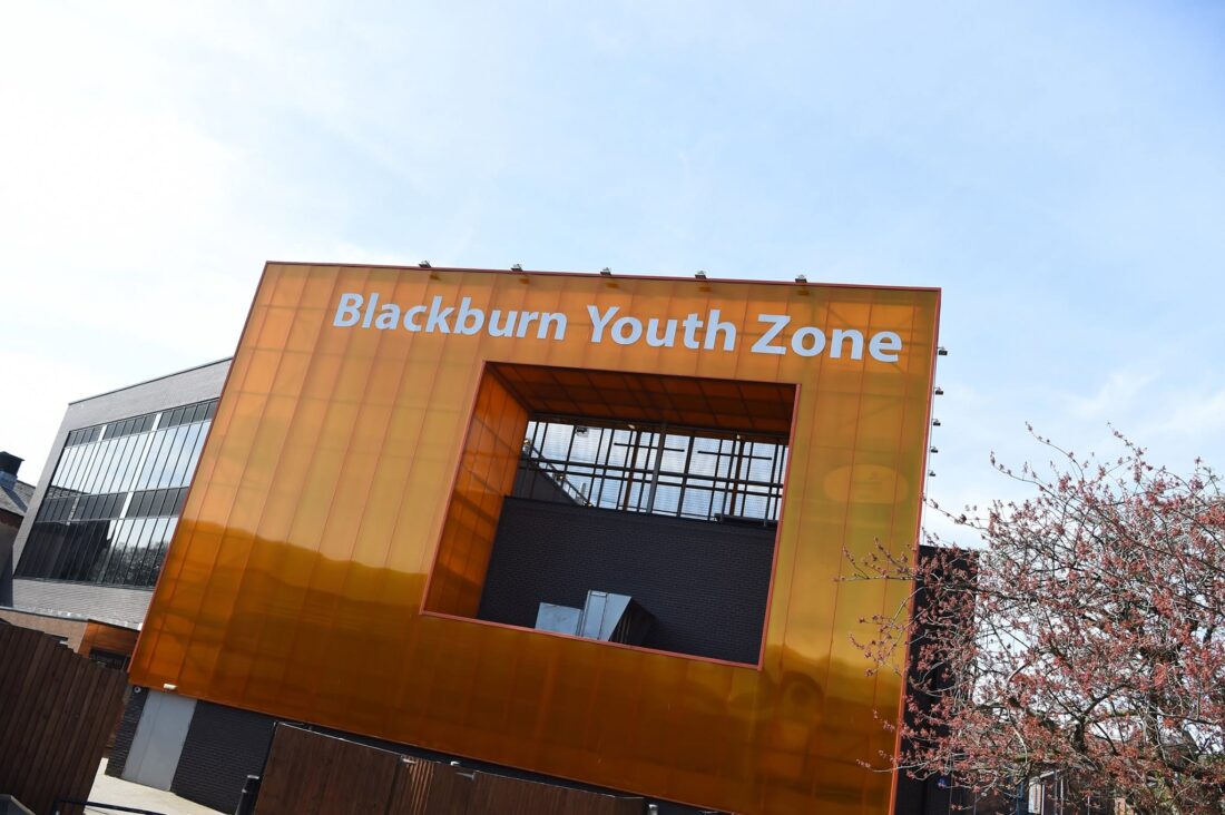The image shows the modern orange building of Blackburn Youth Zone with large white letters. Tree branches are visible on the right under a blue sky.