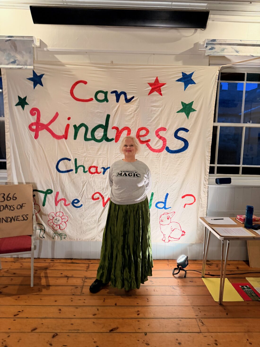 A person stands in front of a banner reading Can Kindness Change The World? with colourful stars. They wear a white shirt and green skirt. A sign on the left says 366 Days of Kindness.