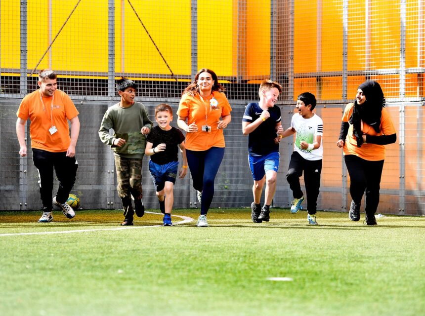Seven people, including adults in orange shirts and children, are running together and smiling on a green field near a metal fence and yellow panels, enjoying an outdoor activity.