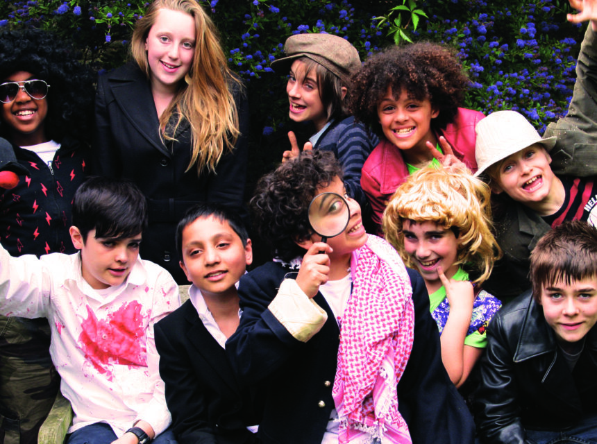 A group of smiling children in fancy dress pose together outdoors, some wearing wigs, hats, glasses, and jackets. One child holds a magnifying glass, and another has a red paint stain on his shirt. Lush greenery is in the background.