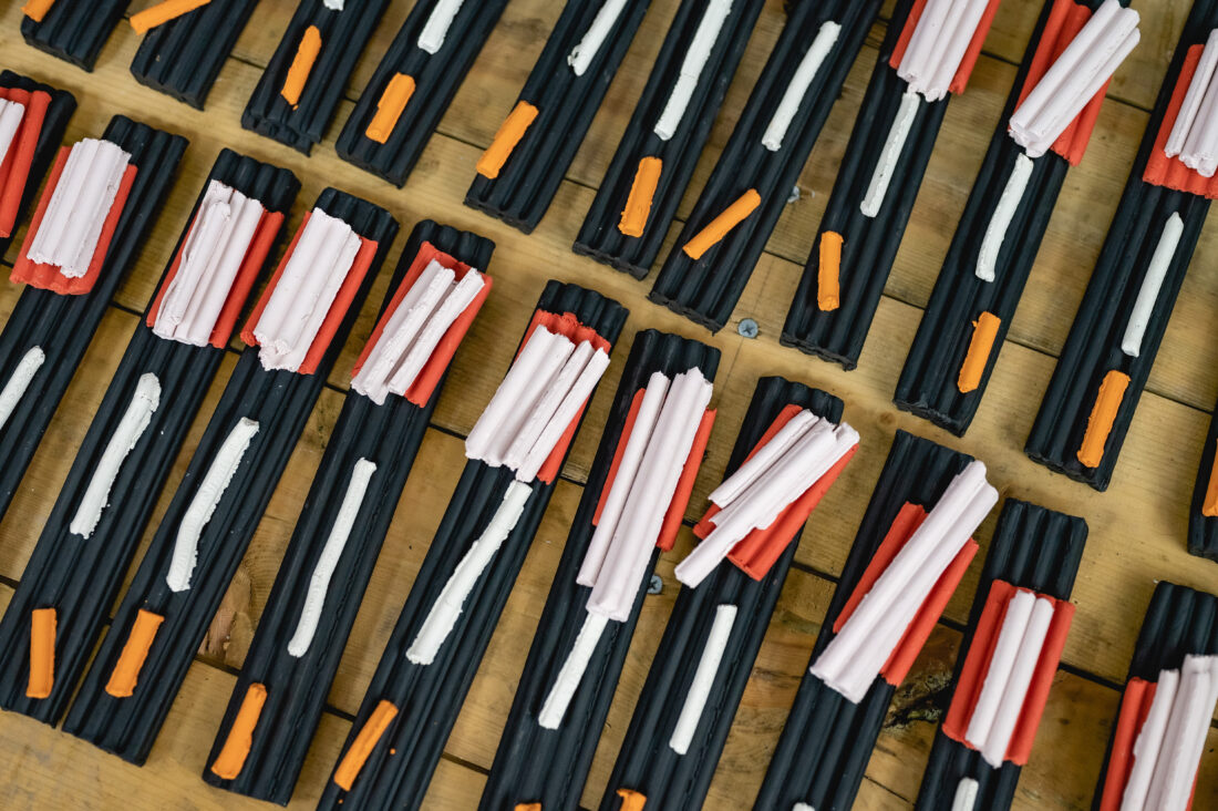 Rectangular black trays arranged on a wooden surface, each holding collections of red, white, orange, and pale pink sticks, possibly sweets or plastic pieces, grouped in varying patterns.