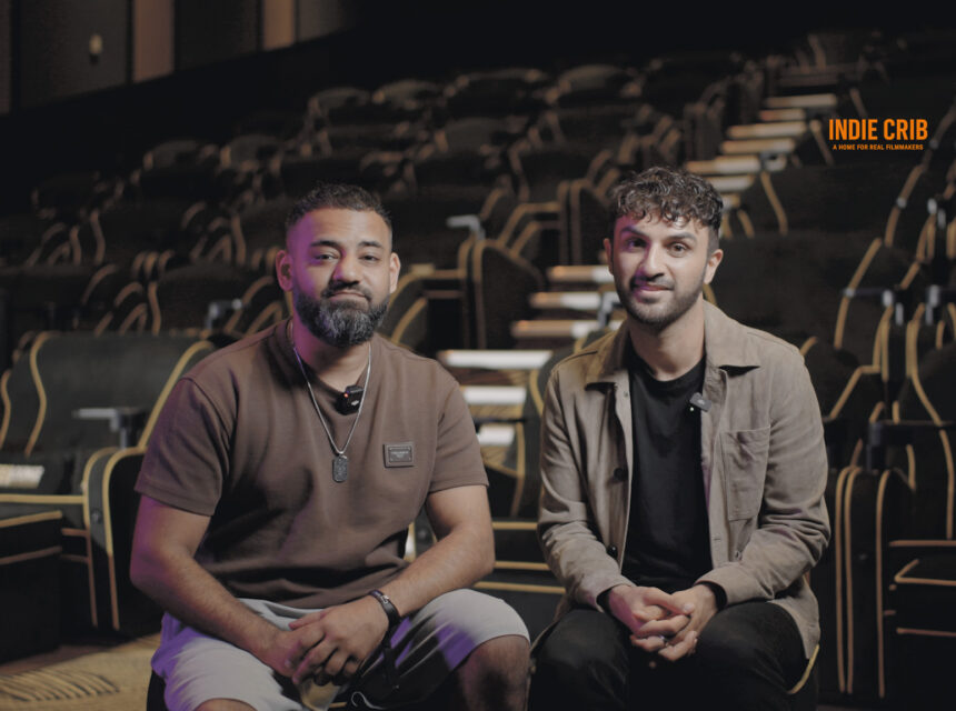 Two men sit in an empty, upmarket theatre with black and gold seats. They face the camera, smiling. A sign reading INDIE CRIB is visible on the wall behind them.