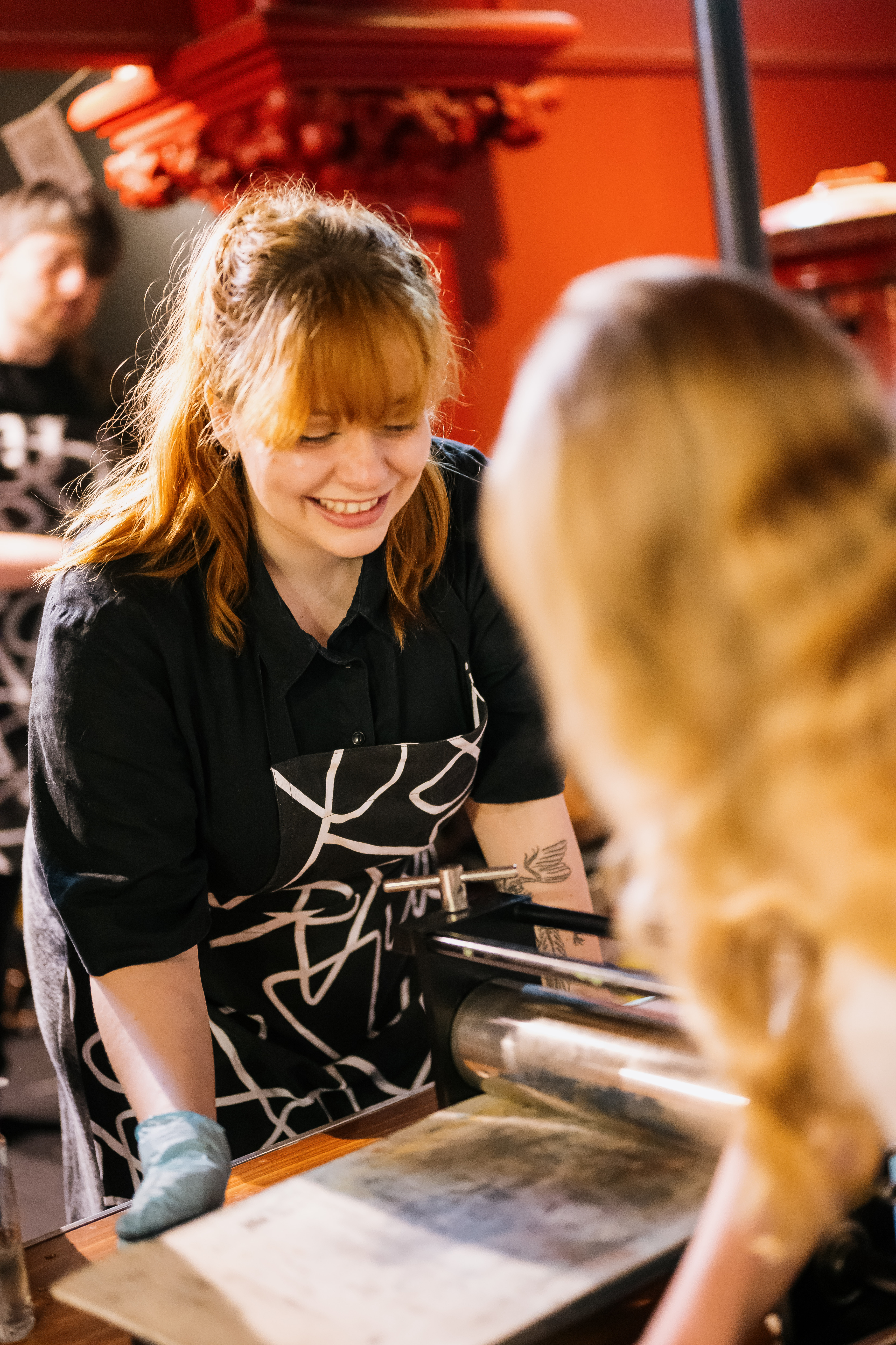 A woman with red hair smiles whilst operating a printmaking press, wearing a patterned apron and glove. Another person stands in the foreground, partially visible. The background is warmly lit with red decor.