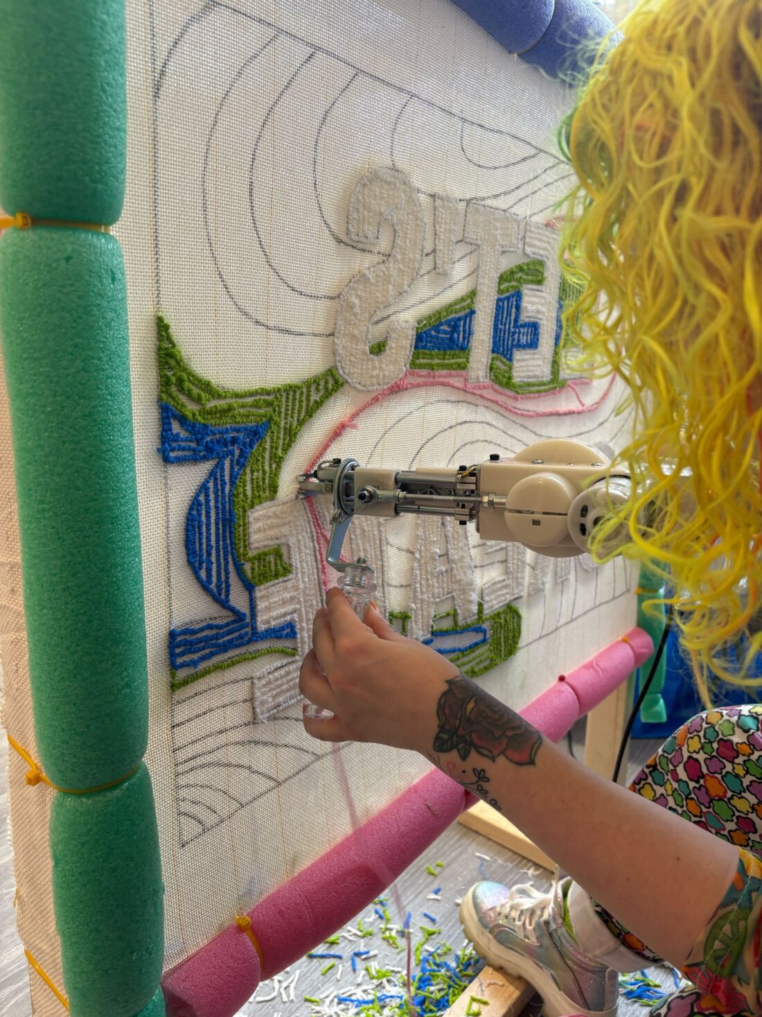 A person with curly yellow hair uses a tufting gun to create a colourful rug design on a large canvas framed with pool noodles. Small pieces of yarn are scattered on the floor.
