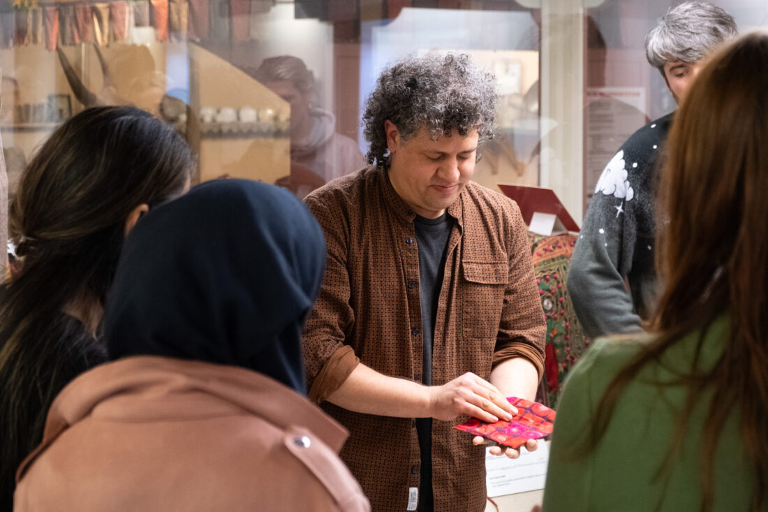 A person with curly hair demonstrates a red and purple patterned object to a small group in a museum setting. Displays and artefacts are visible in the background.