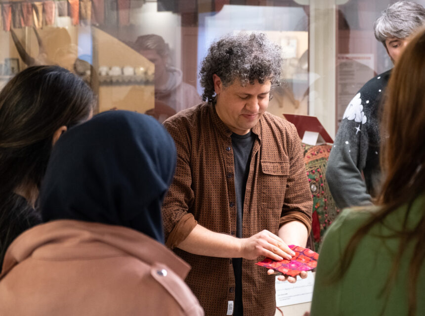 A person with curly hair demonstrates a red and purple patterned object to a small group in a museum setting. Displays and artefacts are visible in the background.