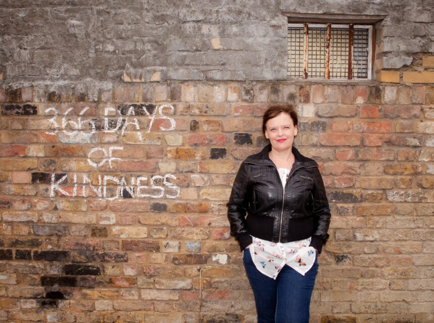 A woman in a black jacket stands against a brick wall with 366 DAYS OF KINDNESS written in white chalk. A small window with bars is at the top of the wall.