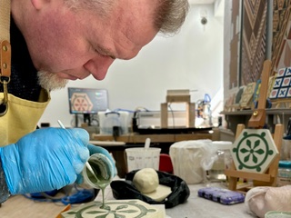 A person wearing blue gloves carefully pours green glaze onto a ceramic tile with a floral pattern in an art studio filled with tools and decorated tiles.