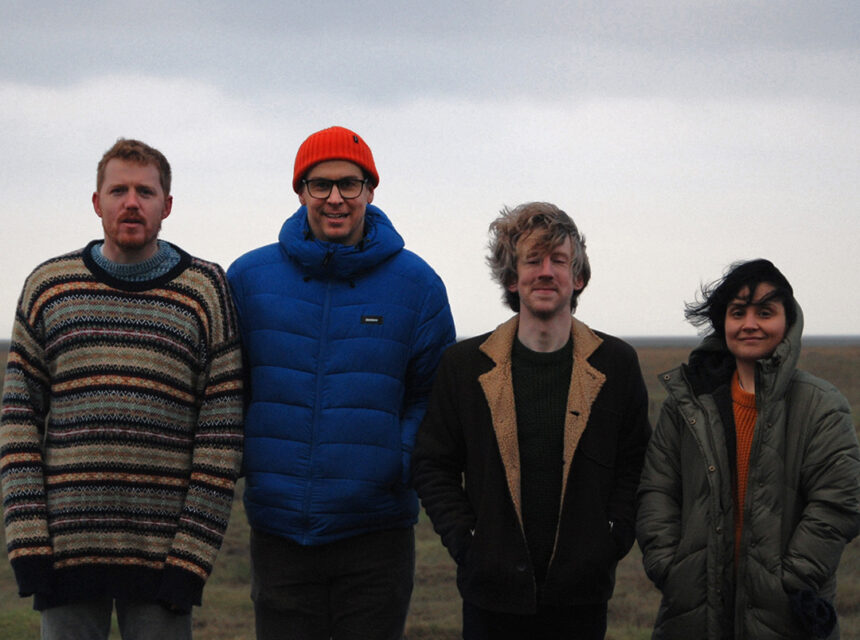 Four people stand side by side outdoors on a cloudy day, all wearing warm coats and jackets, with grass and an overcast sky in the background.