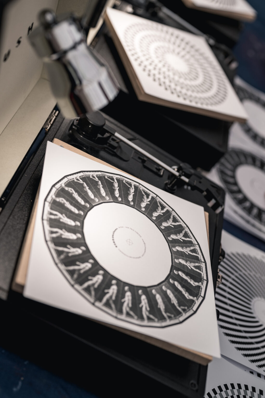 Close-up of printed circular designs with human figures on square sheets placed on record players, resembling old-fashioned animation devices called zoetropes. Other similar patterned sheets are scattered about.