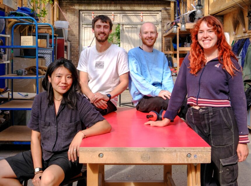 Four people smiling and posing around a red table in a workshop with shelves, jackets, and tools in the background. Sunlight brightens the scene.