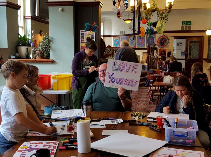 People sit at tables doing crafts in a decorated room. A person in the centre holds a sign that says LOVE YOURSELF. Art supplies and papers are scattered on the tables, and colourful decorations hang from the ceiling.