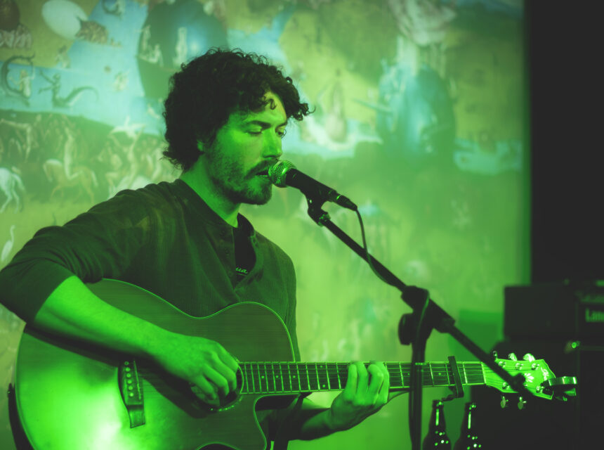 A man with curly hair plays an acoustic guitar and sings into a microphone on stage, bathed in green light, with an artistic mural in the background.