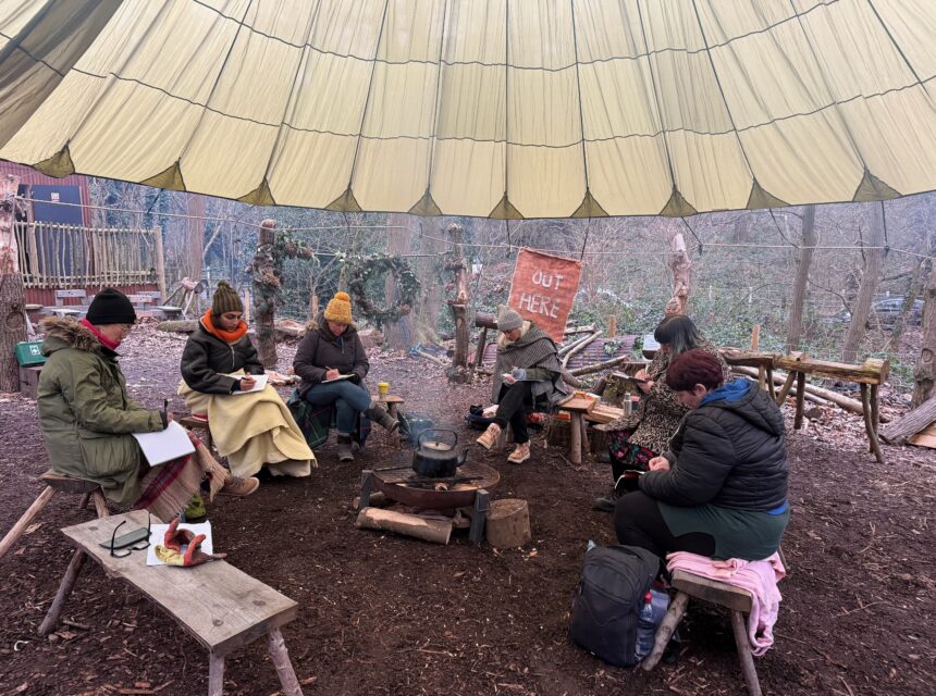 Six people sit around a campfire under a large canopy in a wooded area, writing or drawing in notebooks. Rustic benches and tables surround them, with trees and a hand-painted OUT HERE sign visible in the background.
