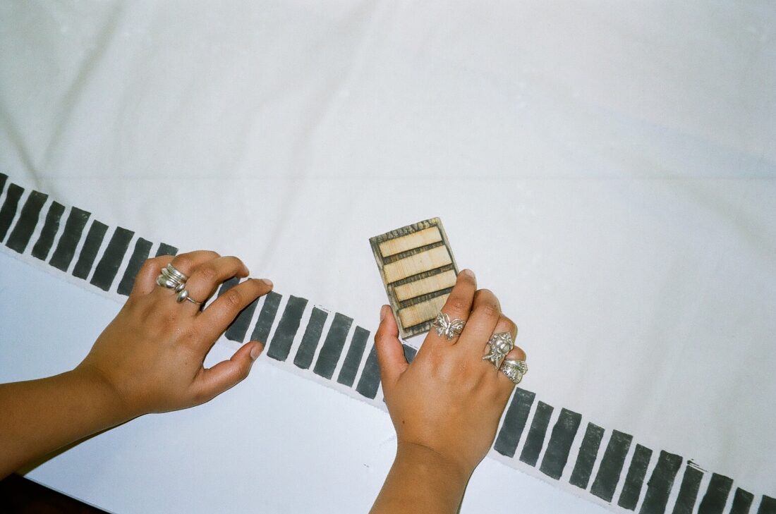 Two hands with several silver rings arrange a small rectangular tool with wooden slats on a white fabric featuring a black geometric border pattern.