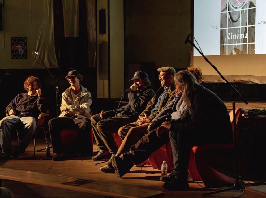 Six people sit on stage in a dimly lit room, engaged in discussion. A microphone and water bottles are visible. A cinema slide is projected on a screen behind them.