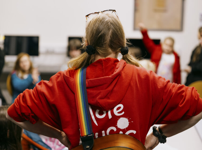A person in a red hoodie with a rainbow strap stands with hands on hips, facing a group of seated children in a classroom setting.