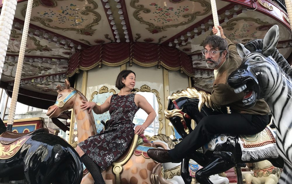 A woman in a patterned dress sits on a giraffe and a man in a brown jumper and glasses sits on a black carousel horse, both smiling while riding the carousel.