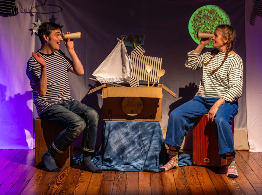 Two people in striped shirts sit on boxes, pretending to look through cardboard tubes. They smile at each other with a homemade boat and moon backdrop between them, on a wooden floor with playful stage lighting.