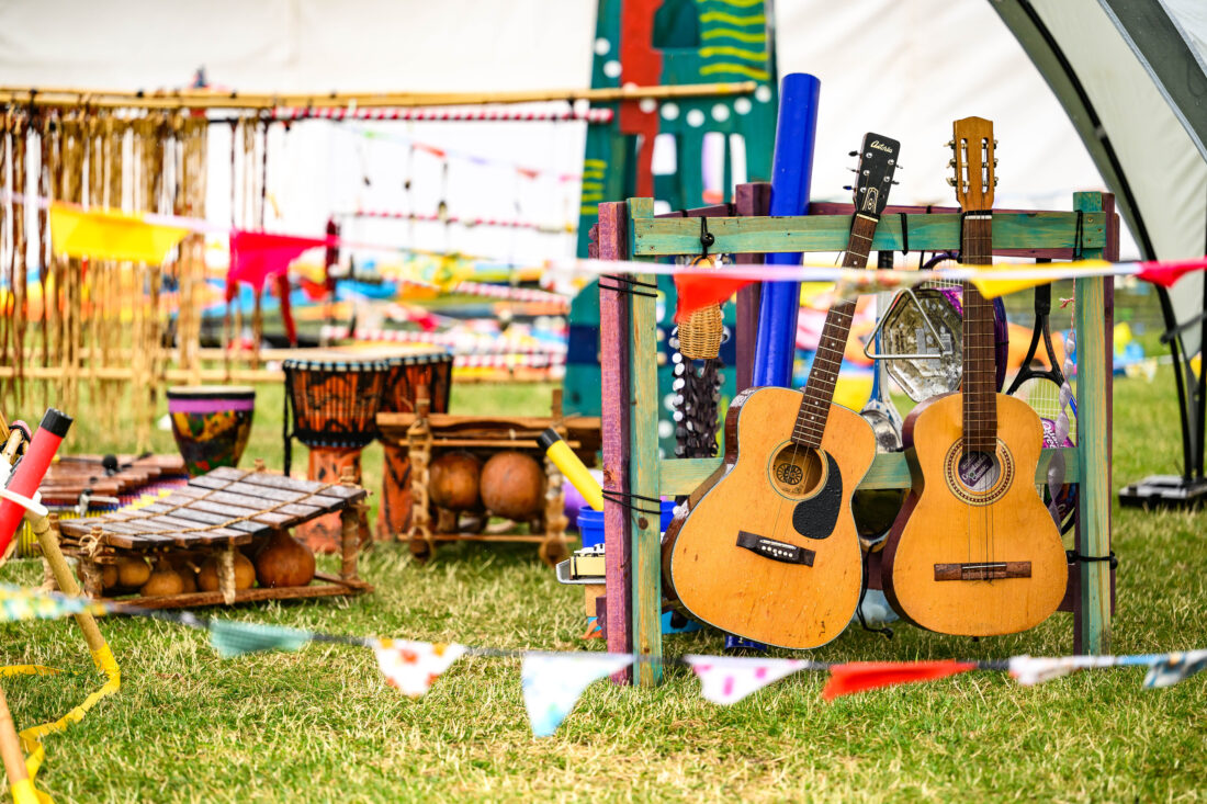 Two acoustic guitars rest on a colourful wooden stand at an outdoor festival, surrounded by musical instruments and vibrant bunting flags, with grass and decorated tents in the background.