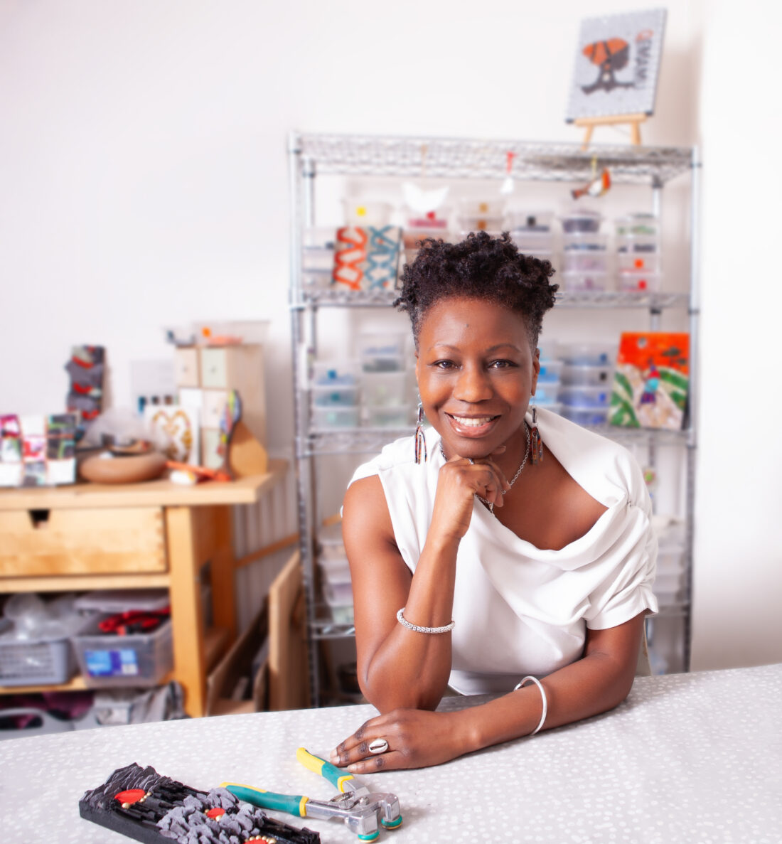 A Black woman with short dark hair and a white shirt smiles at the camera, resting her chin on her hand. She sits at a craft table with tools in front of her. In the background, a metal shelving unit is filled with supplies and art pieces.