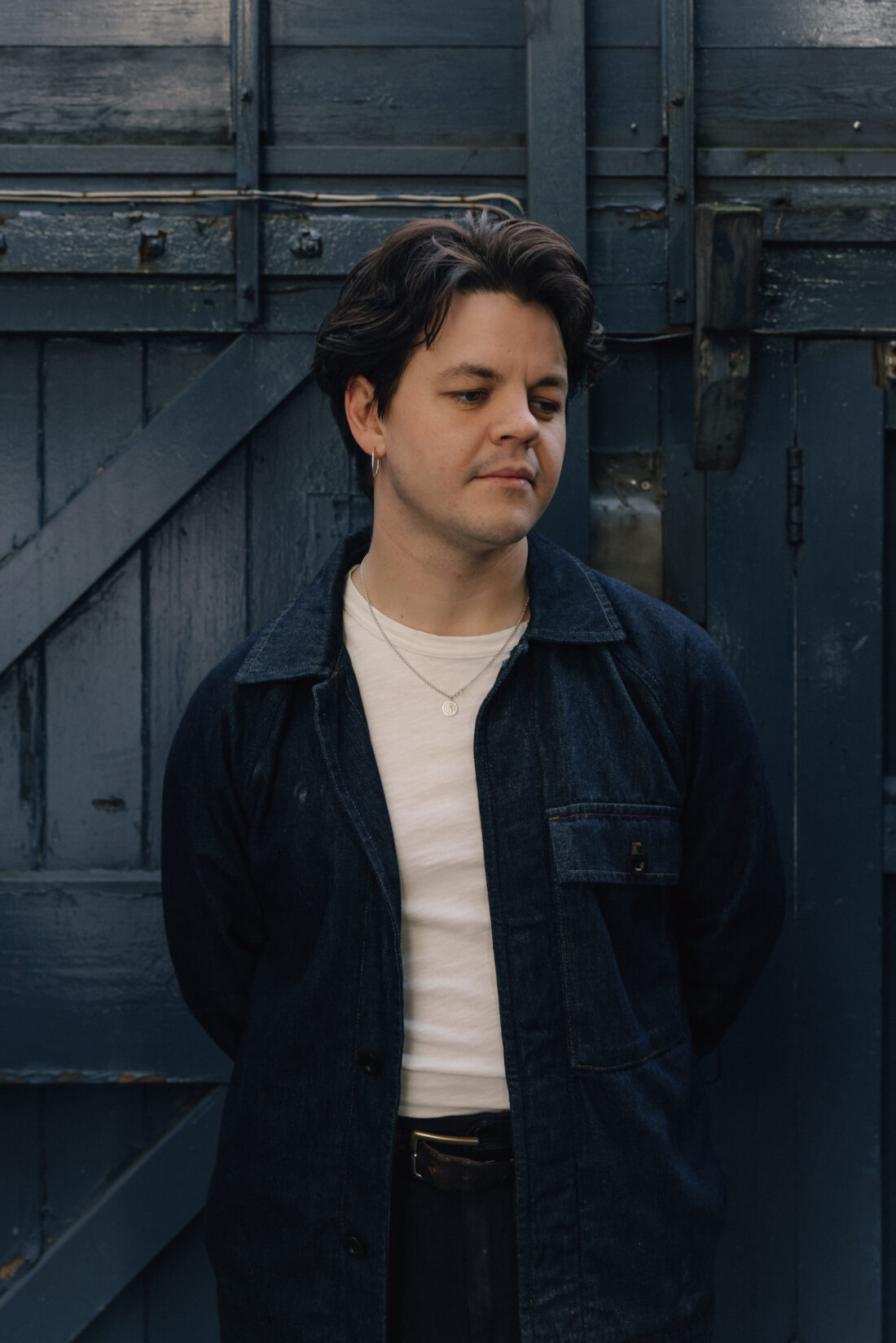 A person with short dark hair, wearing a white shirt and dark jacket, stands in front of a blue wooden door, looking down with hands behind their back.