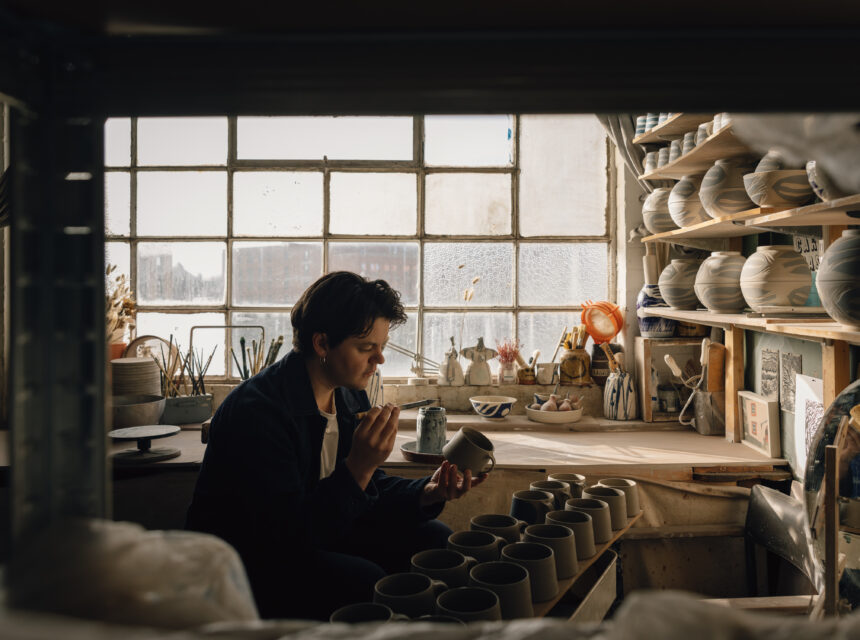 A person sits in a pottery studio, inspecting a mug. Shelves with pottery pieces and tools surround them, and sunlight streams through large windows in the background.
