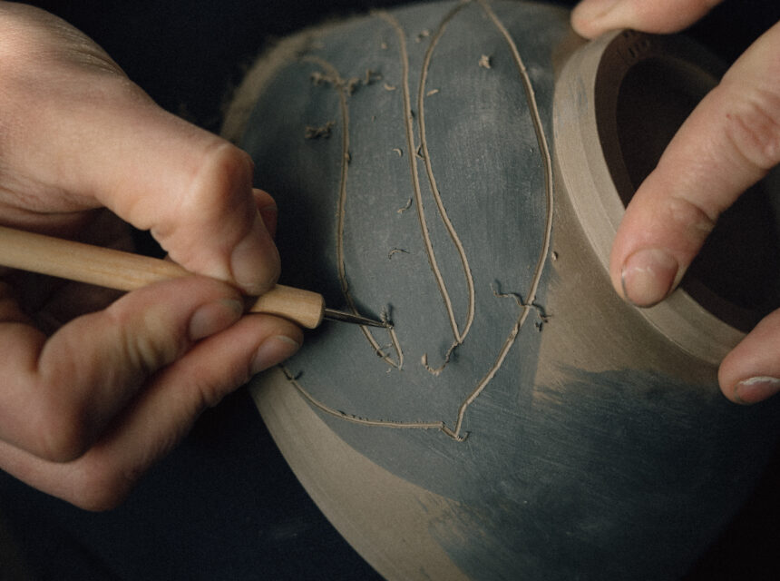 Close-up of a potter’s hands carving a leaf pattern into a dark grey clay bowl using a small wooden-handled tool, with shavings visible on the surface.