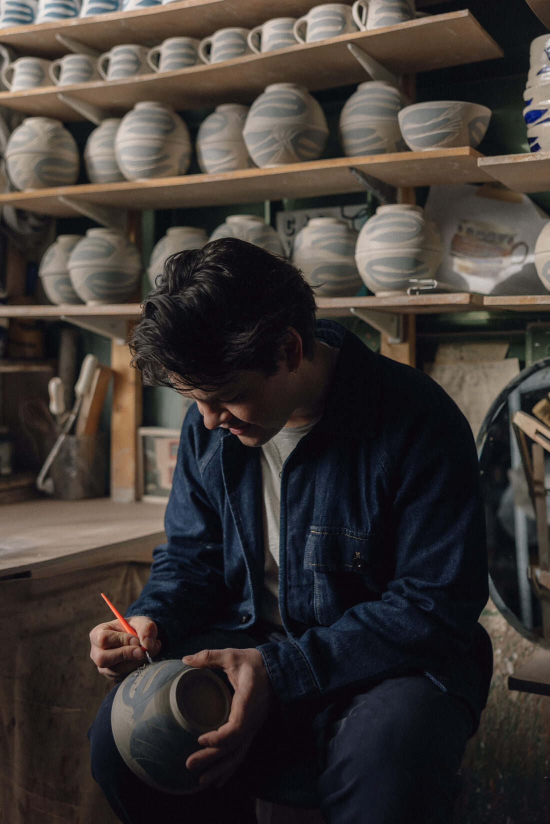 A person sits in a pottery studio, painting designs on a ceramic pot. Shelves behind them display several patterned bowls and cups. The workspace includes brushes and pottery tools.