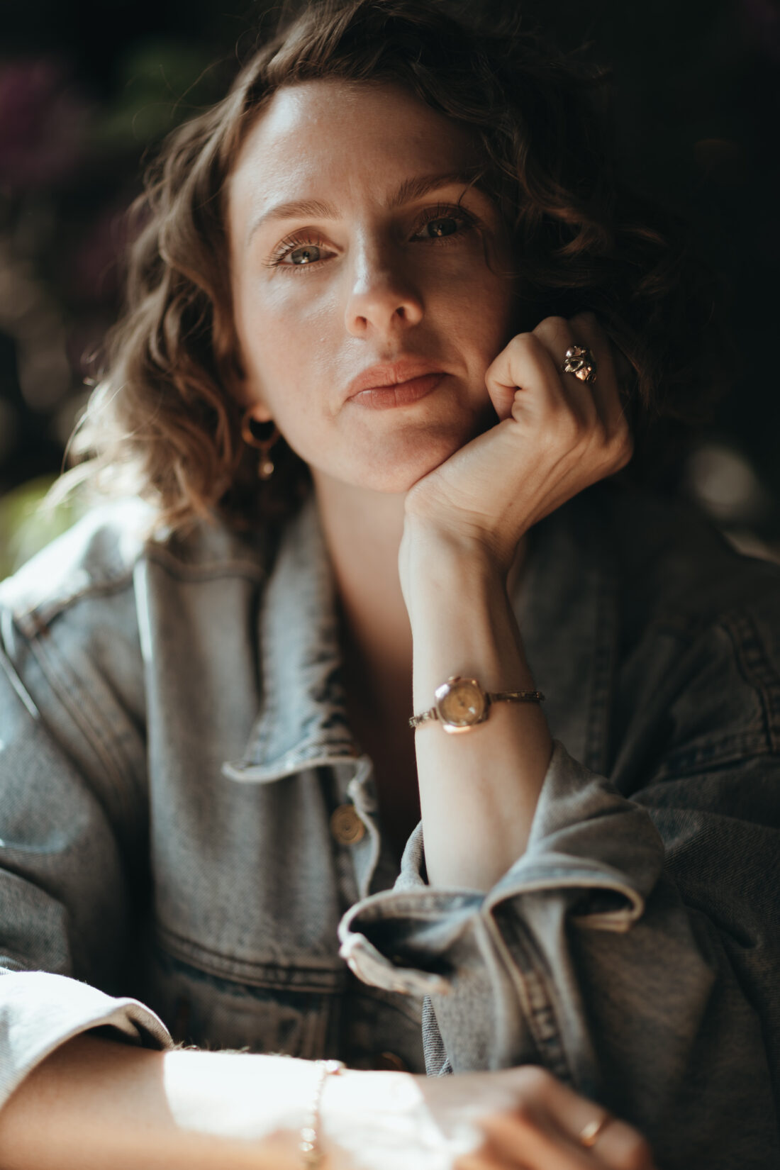 A woman with wavy brown hair rests her chin on her hand, wearing a denim jacket and jewellery, gazing thoughtfully at the camera in soft, natural light.