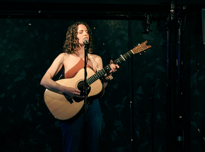 A woman with curly hair plays an acoustic guitar and sings into a microphone on a dimly lit stage with a dark, textured backdrop.