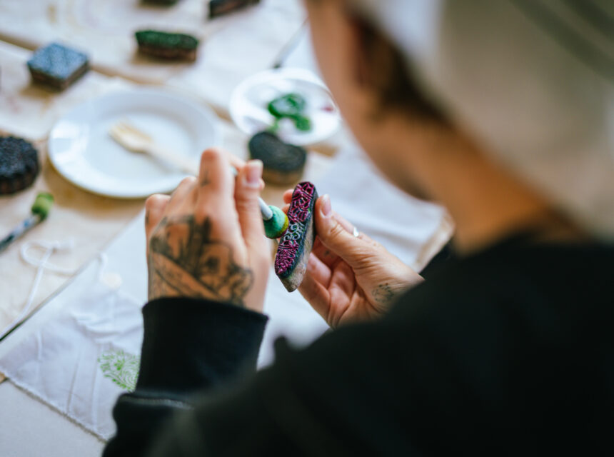 A person with tattoos holds and paints a detailed object, possibly a biscuit or craft, at a table set with art supplies and decorated items. The focus is on their hands and the painted item.