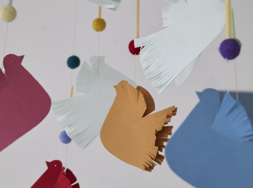 Paper birds in various colours hang from strings along with colourful felt balls, creating a mobile against a soft background. The birds have fringed edges and are suspended at different heights.