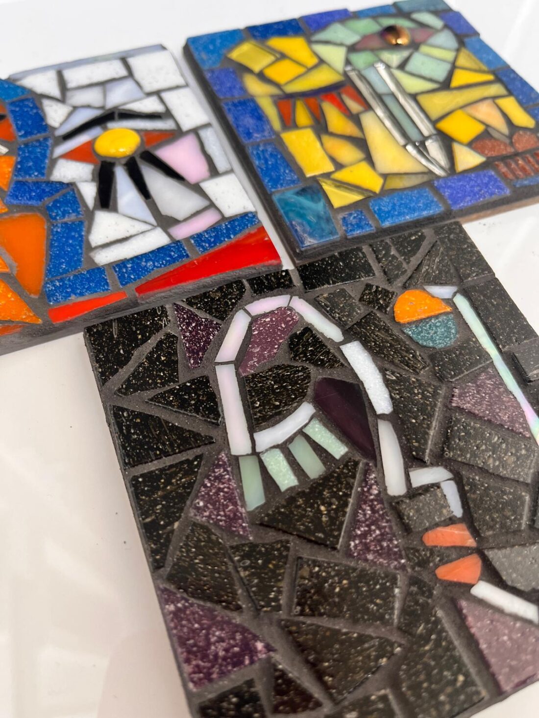 Three colourful, abstract mosaic tiles with various geometric patterns and shapes. The tiles feature vibrant pieces in shades of orange, blue, yellow, black, purple, and white, set against a plain white background.