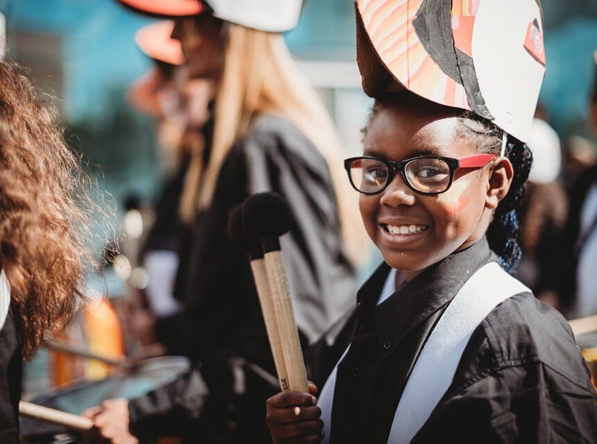 A smiling child with glasses, wearing a colourful hat and holding drumsticks, stands among other children in matching outfits, likely participating in a parade or music event.