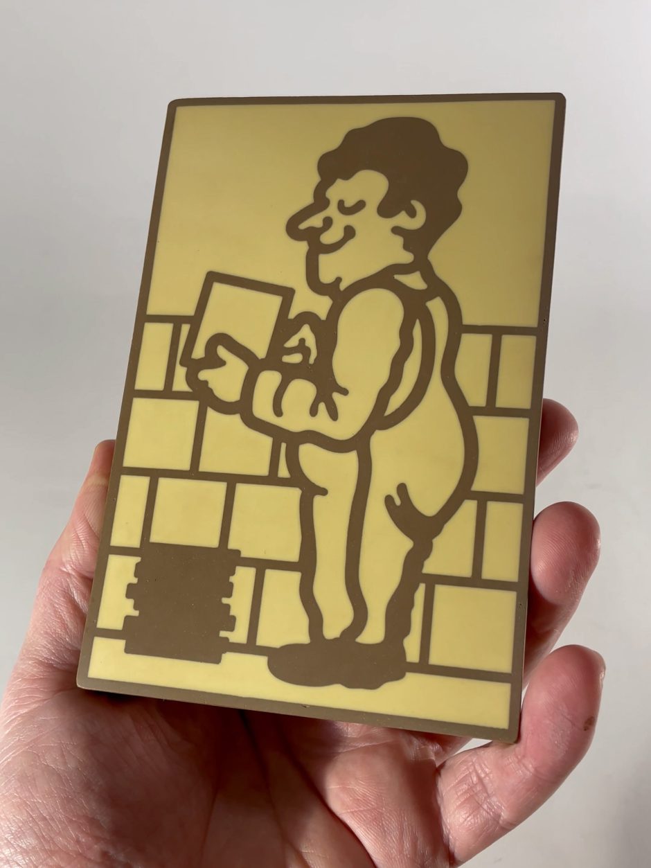 A hand holds a rectangular plaque with an illustration of a smiling person in dungarees building a brick wall. The drawing is simple with brown lines on a yellow background.