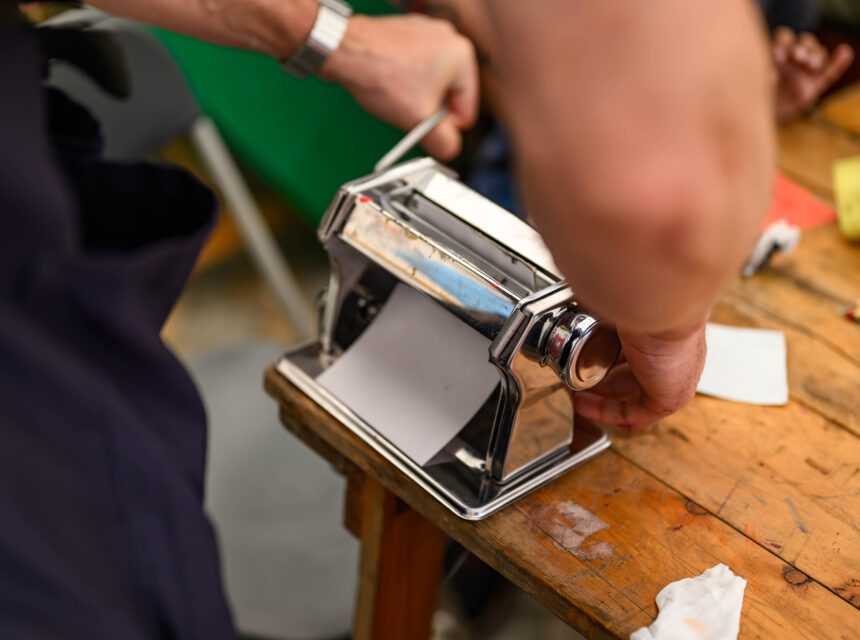 A person uses a metal pasta machine on a wooden table, rolling out dough with one hand while turning the handle with the other. Various kitchen items are visible in the background.