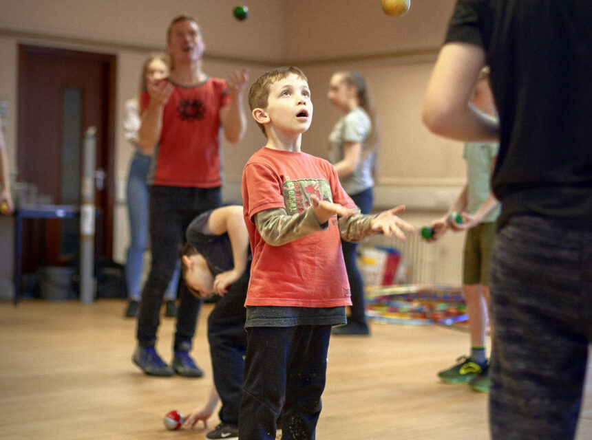 A young boy in a red shirt juggles colourful balls in a room, with other people juggling and watching in the background.