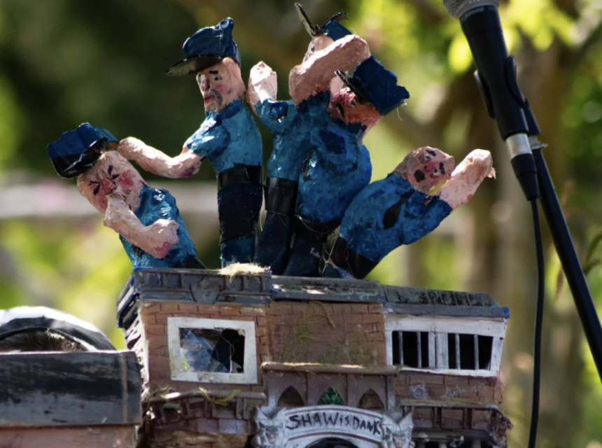 Four blue-uniformed, handmade police officer puppets stand atop a small model building labelled SHANTIS DAYS, next to a microphone, with greenery blurred in the background.