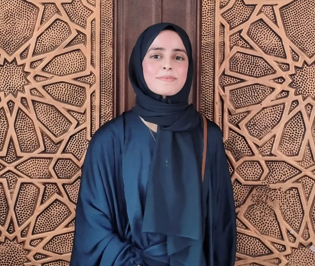 A woman wearing a navy blue hijab and abaya stands in front of an ornate wooden door with intricate geometric patterns.